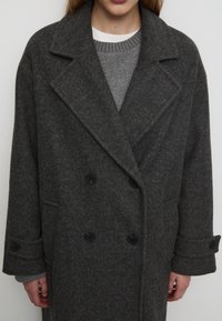 Cappotto grigio in lana doppiopetto con ampi risvolti e bottoni neri. Presenta un motivo a spina di pesce e maniche larghe con risvolto.