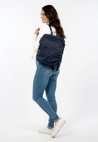 Vrouw in een wit jack en spijkerbroek met een donkerblauwe rugzak, staand opzij en glimlachend tegen een witte achtergrond.