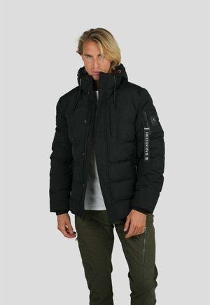 Homme blond portant une doudoune noire avec capuche et étiquette "REDSKINS" sur la manche, pantalon vert olive avec poches zippées et chemise blanche.