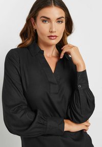 Chemise noire à col, avec un plastron partiel à boutons, des manches bouffantes et des poignets boutonnés. Fabriquée en tissu doux avec une texture lisse.