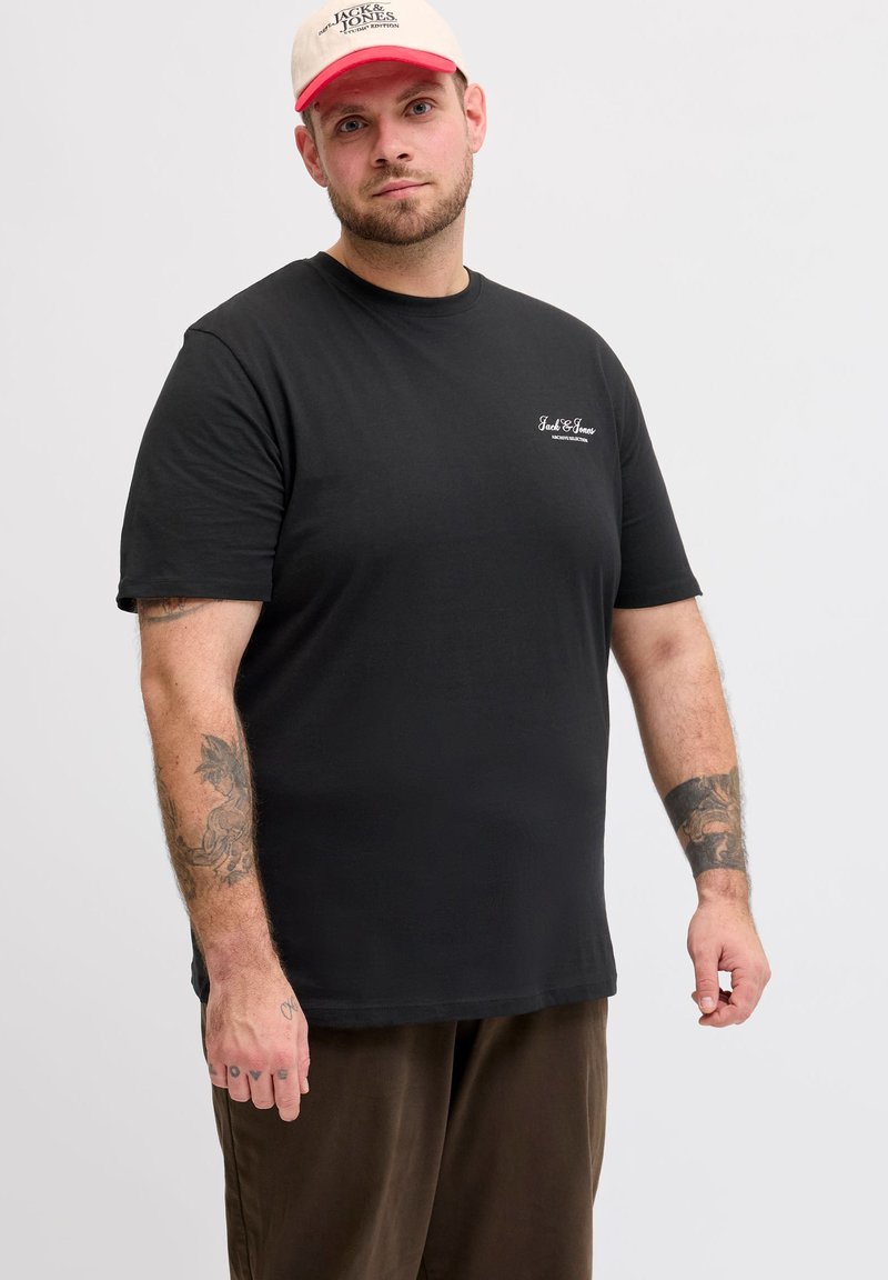 Άνδρας που φοράει μαύρο t-shirt Jack & Jones, καφέ παντελόνι, άσπρο και κόκκινο καπέλο, με ορατά τατουάζ στα χέρια, στέκεται μπροστά σε ένα απλό φωτεινό φόντο.