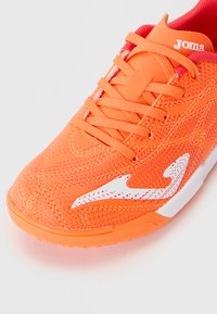 Baskets de sport orange avec lacets, logo incurvé blanc sur le côté, tissu texturé et marque "Joma" sur la languette.