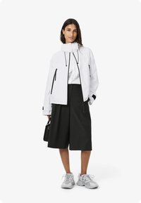 Giacca bianca con zip nere, colletto alto e coulisse, abbinata a una camicetta bianca e pantaloni corti a quadri scuri. Sneakers grigie e borsa nera.