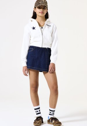 Weißer, taillierter Hoodie mit Sternen-Detail, kombiniert mit einem Jeansrock. Dazu gehört eine Kappe mit Tiermuster, gestreifte Socken und braune, gemusterte Schuhe.