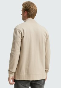 Giacca in jersey beige con maniche lunghe, tessuto testurizzato e vestibilità ampia. Presenta un collo rotondo e un orlo dritto in vita.