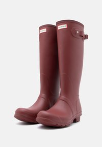 Raudoni guminiai batai su lygiu paviršiumi, apvaliu pirštu, reguliuojamu šoniniu dirželiu ir tekstūruotais padu, užtikrinančiais gerą sukibimą. Sukurta lauko naudojimui.