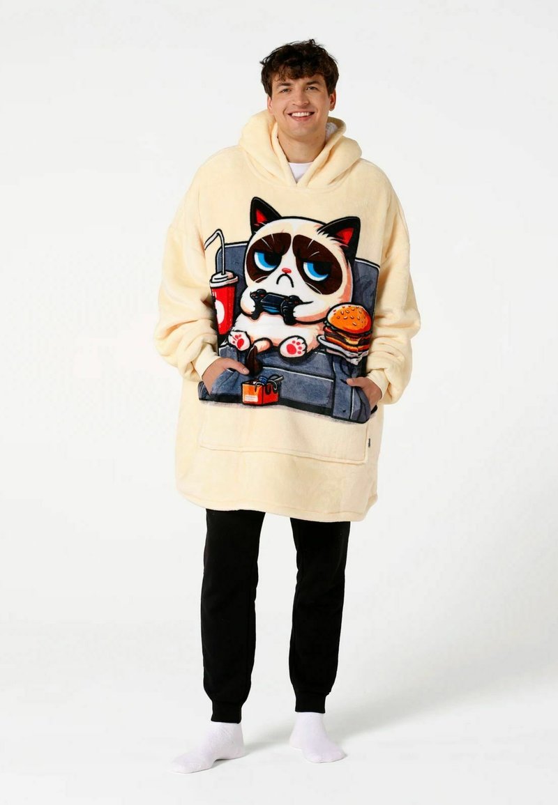 Junger Mann trägt einen übergroßen beigen Hoodie mit einem Cartoon-mürrischen Kater, spielt Videospiele, hält einen Burger und ein Getränk und steht vor weißem Hintergrund.