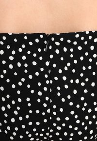 Svart tyg med vita prickar, med dragkedjestängning bak och en slät textur. Designen är lätt och avslappnad.