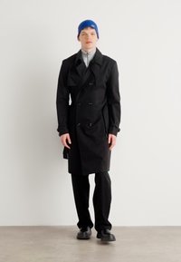 Black double-breasted trench coat with sharp collar, worn over gray turtleneck, paired with black pants and blue knit beanie. Black shoes.