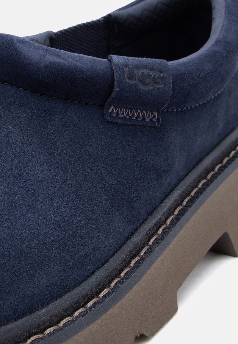 Gros plan sur une chaussure UGG en daim bleu marine avec coutures visibles et semelle marron robuste, mettant en valeur la texture et l'étiquette de la marque.