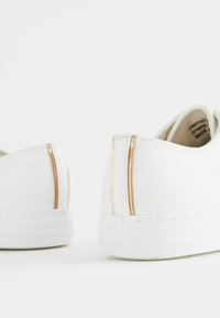 Baskets blanches avec une texture en cuir lisse, dotées de zips dorés subtils au niveau du talon, et d'un design minimaliste avec une semelle plate.