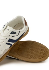 Sneaker bianco con strisce blu navy e un accento in pelle marrone sul tallone. Presenta una suola in gomma testurizzata e fori per i lacci rotondi. Sezione della punta in suede.