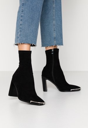 Bottines à talons aiguilles en daim noir avec un bout pointu, ornées d'un détail argenté et d'une fermeture éclair latérale, associées à un pantalon en jean court.