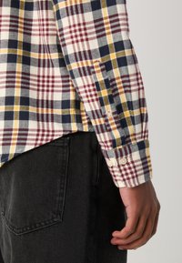 Personne portant une chemise à carreaux à manches longues avec des rayures rouges, jaunes et bleu marine et un pantalon noir, montrant la zone du bras et de la poche latérale.