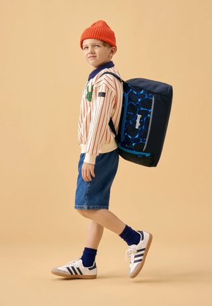 Child in a striped long-sleeve shirt with a green detail, denim shorts, and sneakers, carrying a blue-patterned backpack. Orange knit hat.