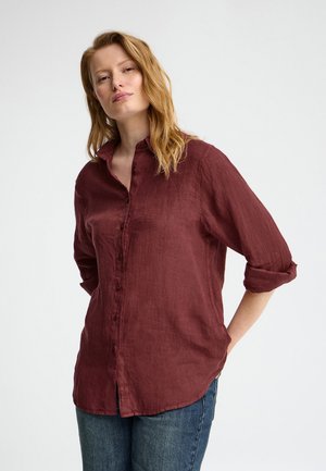 Femme aux cheveux roux ondulés, portant une chemise bordeaux à manches longues avec les manches retroussées et un jean bleu, debout devant un fond uni.