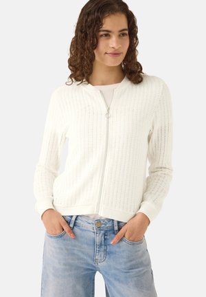 Kvinde iført en hvid tekstureret cardigan med lynlås og lyseblå jeans, der står med hænderne i lommerne foran en ensfarvet baggrund.
