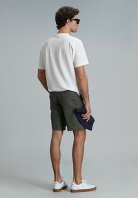 Witte korte mouwen shirt en olijfgroene shorts, met een marineblauwe pet in de hand. Bevat witte sneakers met lichtbruine zolen. Krullend haar.