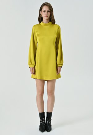 Abito giallo in satin con maniche lunghe e gonfie e colletto alto. Abbinato a stivali neri sopra la caviglia con accenti metallici.