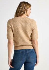 Maglione lavorato a maglia in un morbido beige con maniche a sbuffo corte. Presenta un motivo testurizzato e un'orlo a coste. Abbinato a jeans attillati blu scuro.