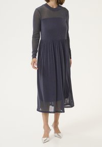 Marinblå långärmad klänning med figursydd kropp och rynkad kjol, tillverkad av ett glittrande genomskinligt material. Har en rundad halsringning.