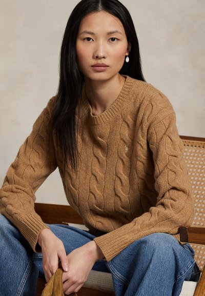 Mladá žena s dlouhými černými vlasy, oblečená v hnědém svetr s copánkovým vzorem a modrých džínách, sedící na dřevěné židli s opěradlem z ratanu.