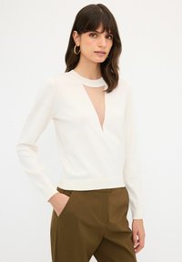 Weißer Langarmpullover mit V-förmigem Ausschnitt am Hals. Weicher Stoff und taillierte Passform. Kombiniert mit braunen hoch taillierten Hosen.