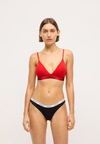 Raudonas medvilninis bralette su plonomis petnešėlėmis ir juodu Calvin Klein logotipo elastiku, derinamas su juodu nailono thong'u su baltu logotipo juostele.