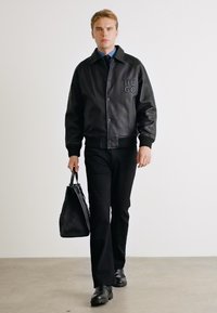 Melna ādas bomber jakas ar ribotām piedurknēm, kurai ir iespiests "HUGO" zīmols. Modelis valkā melnas bikses un nes teksturētu melnu somu.