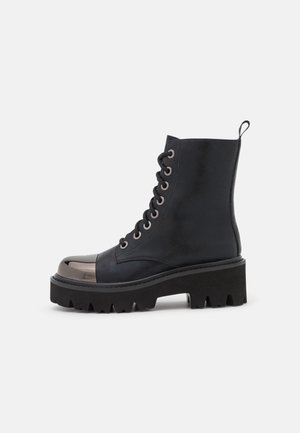 Bota negra de tacón bajo con cordones, suela gruesa con dibujo pronunciado y puntera metálica brillante, vista lateral sobre fondo blanco.