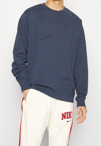 Man som bär en marinblå Nike-tröja och vita Nike-träningsbyxor med röda ränder och logotyper, med handen i fickan, står framför en enfärgad bakgrund.