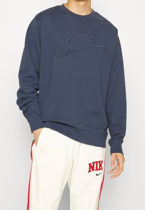 Uomo che indossa una felpa Nike blu navy e pantaloni della tuta Nike bianchi con strisce e loghi rossi, con una mano in tasca, in piedi davanti a uno sfondo neutro.