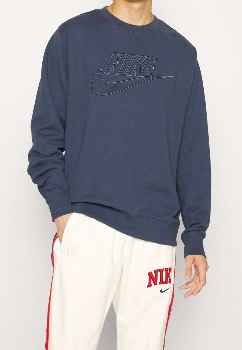 Man som bär en marinblå Nike-tröja och vita Nike-träningsbyxor med röda ränder och logotyper, med handen i fickan, står framför en enfärgad bakgrund.