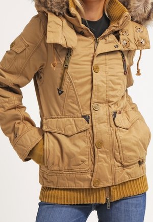 Donna che indossa una giacca invernale beige con cappuccio e rifiniture in pelliccia, vari tasche, bottoni e cerniere, abbinata a jeans blu e maglione senape.