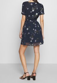 Žena v námořnickomodrých květinových šatech s krátkým rukávem a elastickým páskem, v černých lodičkách, stojící na jednobarevném pozadí.