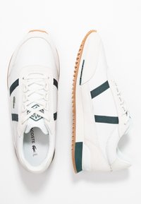 Baskets blanches en cuir texturé, avec des rayures vert foncé, une semelle extérieure en gomme, et un petit logo Lacoste sur le côté.