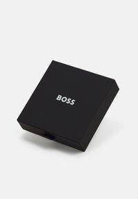 Cutie neagră dreptunghiulară cu o suprafață texturată, având un logo argintiu "BOSS" în față și un mic tab pentru deschidere.