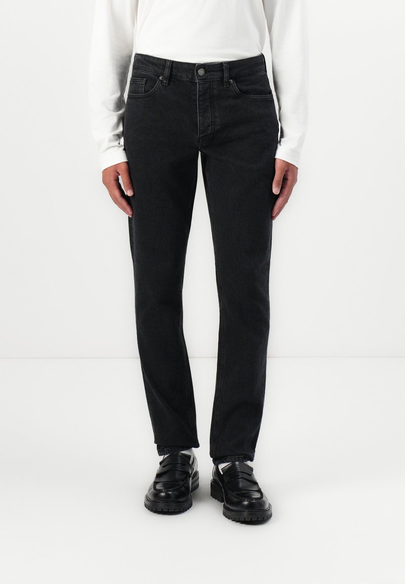 Fekete farmernadrág slim fit fazonban, öt zsebbel, övhurkokkal és gombos zárással. Hosszú ujjú fehér inggel és fekete cipővel viselve.