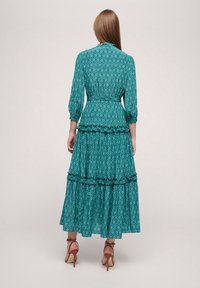 Robe maxi à manches longues en teal avec des motifs en losange, taille cintrée et couches en dentelle. Associée à des chaussures à talons hauts marron clair.
