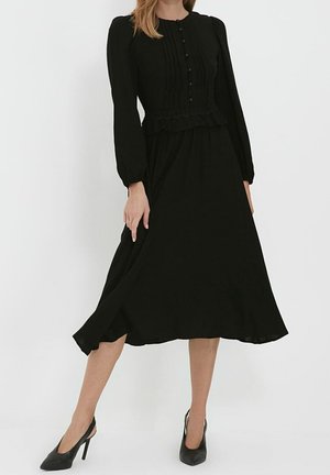 Kvinde iført en sort midi-kjole med lange ærmer med knapdetaljer og plisseringer, kombineret med sorte højhælede slingback-sko.