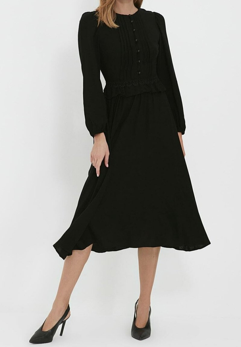Femme portant une robe midi noire à manches longues avec des détails de boutons et des plis, associée à des chaussures slingback noires à talons hauts.