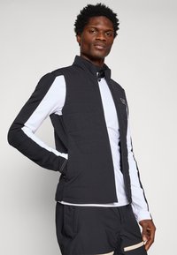 Mustmustatud tikandiga vesti kõrge kraega ja koos lukuga, kombineeritud valge pika varrukaga topiga. Omab külgmisi taskuid ja kontrastsest niidist õmblusi.