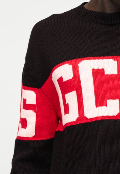 Πλεκτό πουλόβερ με μεγάλα κόκκινα και λευκά γράμματα "GCS" στο στήθος και το πάνω μέρος του μπράτσου, σε μαύρο φόντο, που το φοράει ένα άτομο.