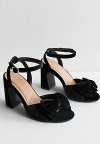 Zwarte fluwelen hoge hak sandalen met een strikdetail, open teen ontwerp, enkelbandjes en een stevige hak. Gladde textuur en neutrale binnenzool.