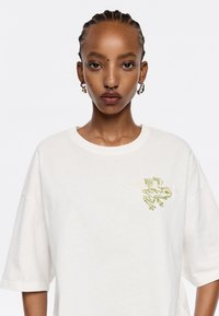 Mujer con cabello trenzado, lleva una camiseta blanca con una rana alada bordada en verde en el pecho y pendientes de aro.