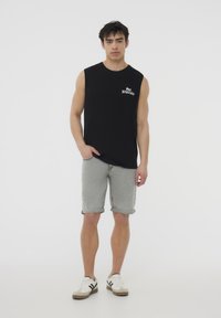 Maglietta nera senza maniche con testo "San Fracticed", abbinata a pantaloni corti in denim grigio chiaro arrotolati all'orlo. Sneakers bianche e grigie completano il look.