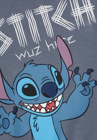 T-shirt gris avec un personnage de dessin animé bleu, aux grands yeux et au sourire ouvert, le texte indique "STITCH WUZ HERE" en blanc.