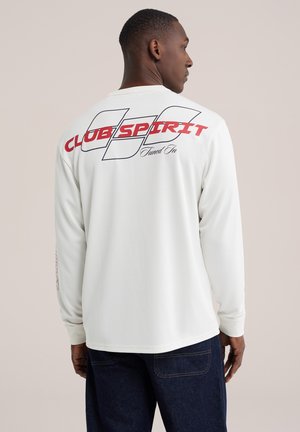 Homme portant une chemise blanche à manches longues avec un texte "CLUB SPIRIT Tuned In" rouge et noir au dos et un jean bleu foncé, de dos.