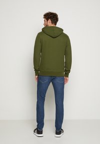 Olivgrüner Kapuzenpullover mit Zugkordel, gerippten Bündchen und Saum; kombiniert mit blauen Jeans und schwarzen Sneakers. Glatte Textur, lässige Passform.