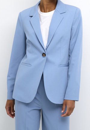 Kvinde iført en lyseblå skræddersyet blazer med én knap over en hvid t-shirt, kombineret med matchende lyseblå bukser.